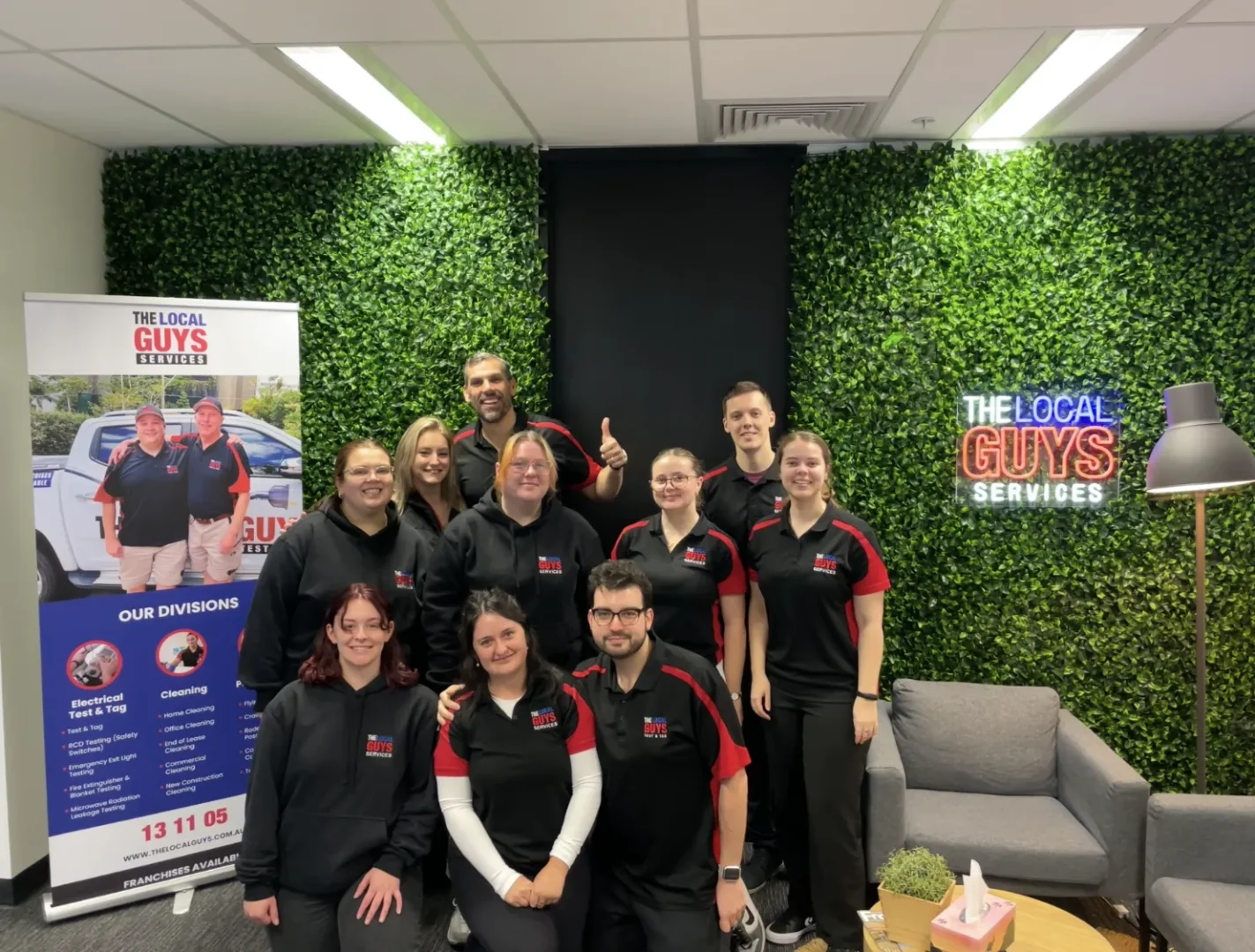 Team of The Local Guys Pest Control in branded attire, posing in a modern office setting with a green wall, showcasing a banner highlighting their services and franchise opportunities.